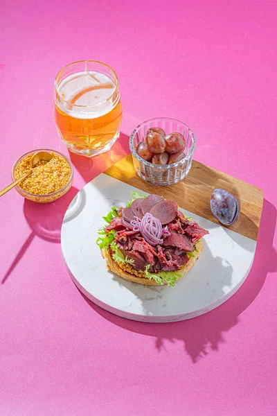 Hamburguesa de pastrami con mostaza, lechuga, pepinillos y cebolla sobre un pan de sésamo recién horneado sobre fondo rosa — Foto de Stock