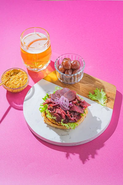 Hamburguesa de pastrami con mostaza, lechuga, pepinillos y cebolla sobre un pan de sésamo recién horneado sobre fondo rosa — Foto de Stock