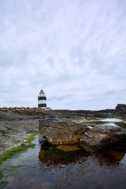 Ön planda kayalar ve bulutlu gökyüzü olan eski Kanca deniz feneri. Wexford, İrlanda.