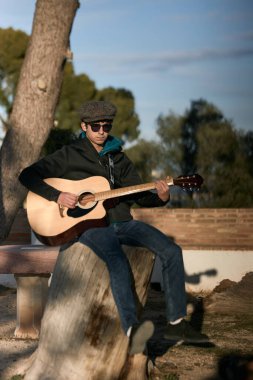 Genç adam çekici bir kıyafetle dışarıda gitar çalıyor..