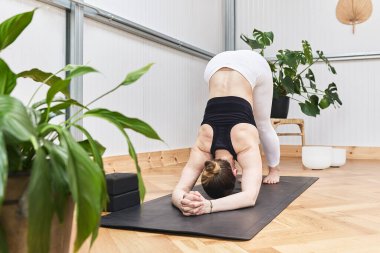 Beyaz kadın yeşil bitkilerle çevrili bir spor salonunda yoga yapıyor.
