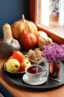 Pencere kenarında farklı türde balkabaklarının yanı sıra bir fincan siyah çay, kabak çekirdeği, badem ve susamlı kurabiyeler ve siyah tepside elma ve hurma dolu bir tabak bulunur. Yakın çekim.