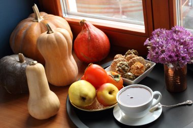 Pencere kenarında farklı türde balkabaklarının yanı sıra bir fincan siyah çay, kabak çekirdeği, badem ve susamlı kurabiyeler ve siyah tepside elma ve hurma dolu bir tabak bulunur. Yakın çekim.