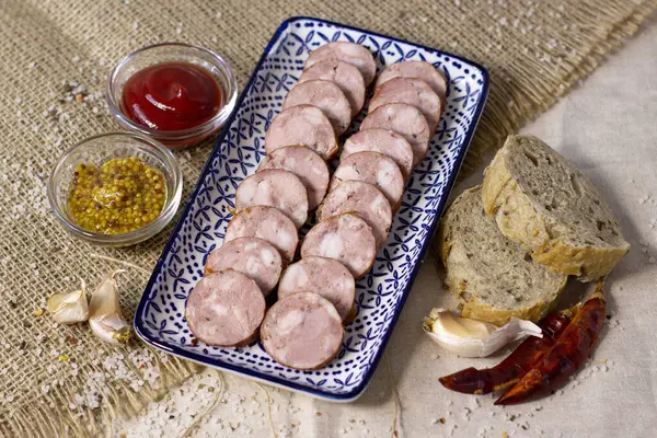 Аппетитная колбаса, нарезанная на синей тарелке на грубом льняном фоне с перцем чили и специями, закрывается — стоковое фото