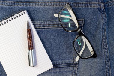 Siyah beyaz çerçeveli şık gözlükler, kot pantolon arka planında not defteri ve kalem dar odak çizgisi, sığ alan derinliği