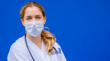 Mavi arka planda koruyucu maskeyle çalışmaya hazır genç ve güzel bir hemşireyi kameraya doğru çek.