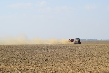 Tractor rides on the field and makes the fertilizer into the soil. Clouds of dust from the dry soil tractor trailer. Fertilizers after plowing the field. clipart