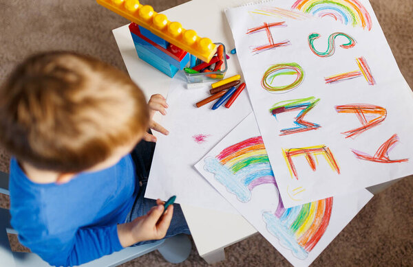 Pandemic coronavirus slogan Stay home. Caucasian boy draws house and word stay home on paper with pencil. Call to self-isolate in event of coronavirus pandemic and not leave house on street.Top view
