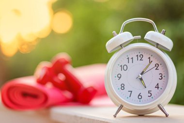 Alarm çalıştırmak için saat 5 'te saat, yeşil arka planda bulanık halterler ve yoga minderi güneş ışığı, spor ve egzersiz konsepti ile dışarıda.