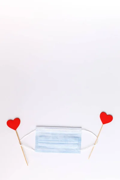 Two red hearts and blue face protection mask in middle, white background. Disposable medical mask and two hearts. Appreciation to medical staff. Valentine Day concept in new reality 2021