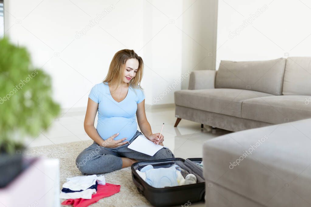 Pregnant woman in blue t-shirt ,checklist and preparing stuff for maternity hospital at home. Happy pregnancy packing baby clothes on suitcase