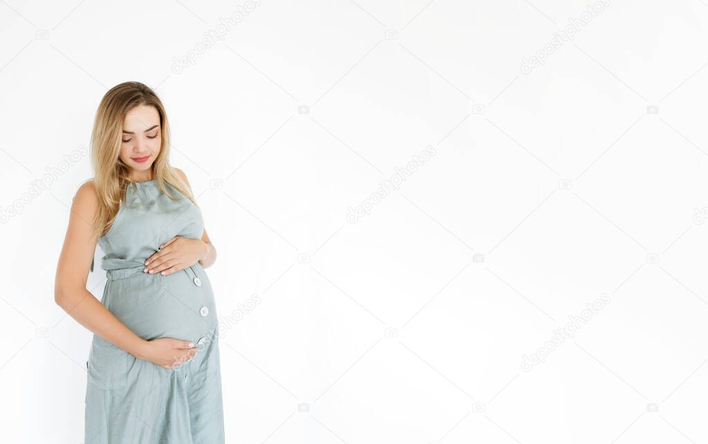 Young pregnant woman holds her hands on her swollen belly in dress. Love concept. Horizontal with copy space, white wall