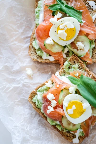 Open smorrebrod sandwich salmon, cucumber, boiled egg, soft cheese. Homemade sandwich with rye bread on white background, top view. Tasty salmon fish sandwich closeup