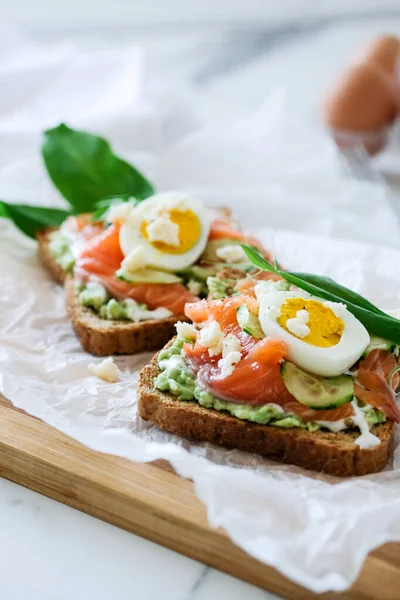 Open smorrebrod sandwich salmon, cucumber, boiled egg, soft cheese. Homemade sandwich with rye bread on white background, top view. Tasty salmon fish sandwich closeup