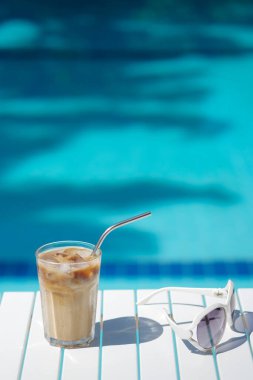 Kıbrıs Rum Kesimi buzlu kahve Frappe Fredo yüzme havuzunun açık mavi suyuna karşı, beyaz masada, güneş gözlüklü. Yaz minimalist geçmişi, tatil veya tatil kavramı. Boşluğu kopyala