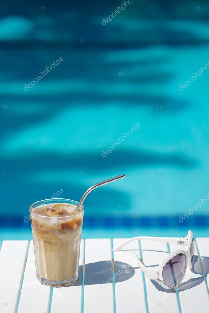 Ice coffee Cyprus Frappe Fredo against blue clear water of the swimming pool, on white table, with sunglasses . Summer minimalistic background, holiday or vacation concept.Copy space