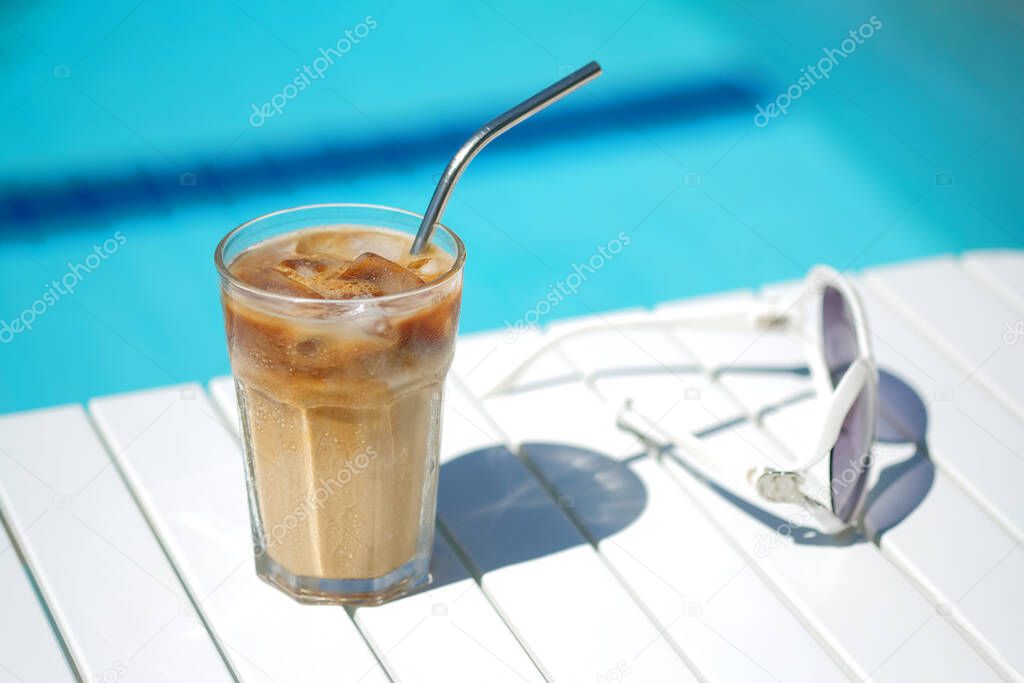 Ice coffee Cyprus Frappe Fredo against blue clear water of the swimming pool, on white table, with sunglasses . Summer minimalistic background, holiday or vacation concept.Copy space