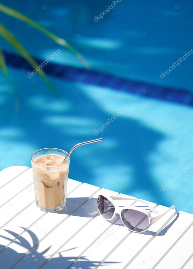 Ice coffee Cyprus Frappe Fredo against blue clear water of the swimming pool, on white table, with metal straw . Summer minimalistic background, holiday or vacation concept.Copy space