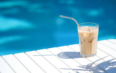 Ice coffee Cyprus Frappe Fredo against blue clear water of the swimming pool, on white table, with metal straw . Summer minimalistic background, holiday or vacation concept.Copy space clipart
