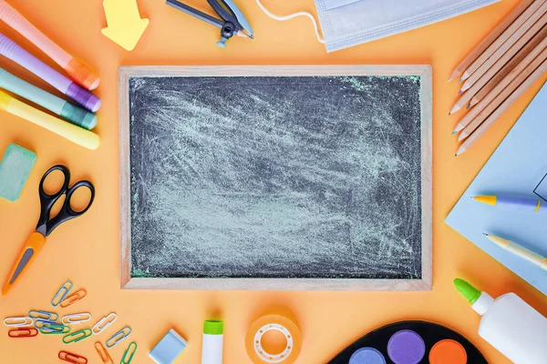 Zurück zur Schule mit Schulmaterial, Schreibwaren, Arztmaske und Notizbuch mit Leerraum in der Mitte, für Kinder auf orangefarbenem Schreibtischhintergrund, von oben flach liegend. — Stockfoto