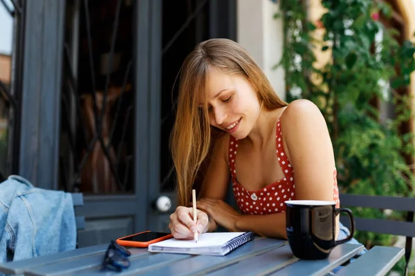 Giovane donna sulla terrazza del caffè. Hipster elegante ragazza concentrata Scrivere in notebook. Tecnologie digitali per lavorare a distanza o studiare in luoghi confortevoli, bere caffè o tè — Foto Stock