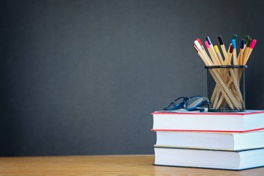 Bir yığın kitap, gözlük, siyah bir okul yönetim kurulunun ahşap arka planındaki metal tutacağında bir sürü kalem. Eğitim kavramı. Boşluğu kopyala.