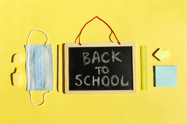 Material de papelaria escolar, máscara médica em fundo amarelo distanciamento social, reabertura da escola após a pandemia de covid-19. Novo conceito normal.Topo espaço de cópia vista, quadro-negro — Fotografia de Stock