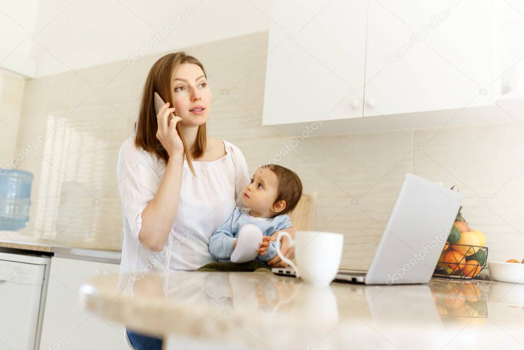 Beautiful business mom is talking on the mobile phone and taking notes while spending time with her cute baby boy at kitchen home office. Working mother concept,Laptop on table.