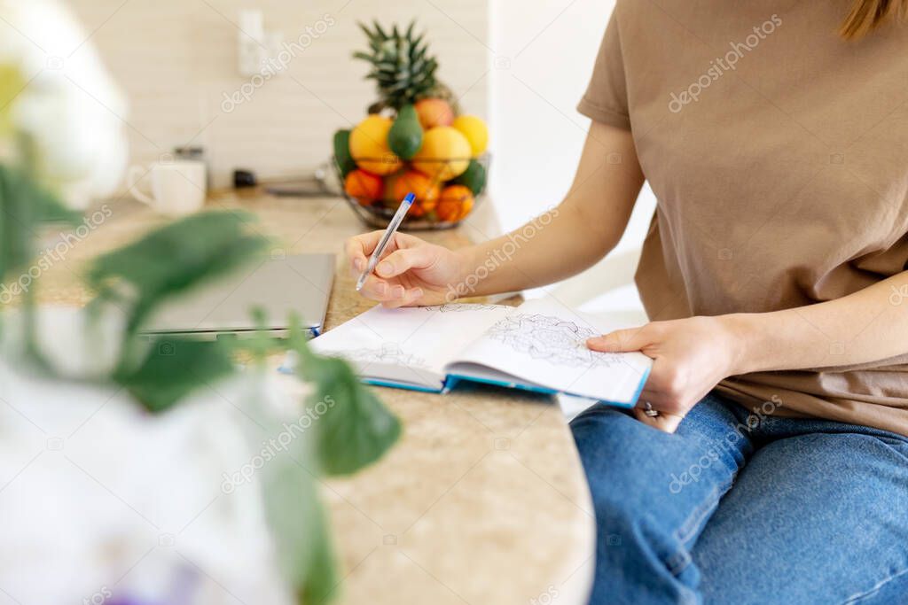 unrecognizable cropped young woman is sitting at the table in her kitchen and writing in a notebook. Making a home shopping list or wish list or to do list.close up.