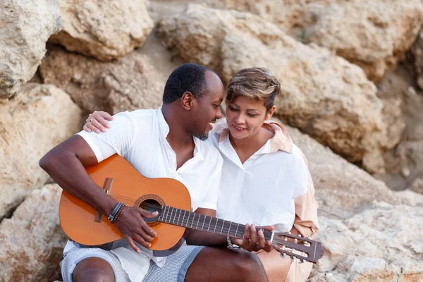 Feliz raça mista, casal de meia-idade abraçando enquanto caminhava na praia segurando uma guitarra. Homem segurando guitarra com mulher — Fotografia de Stock