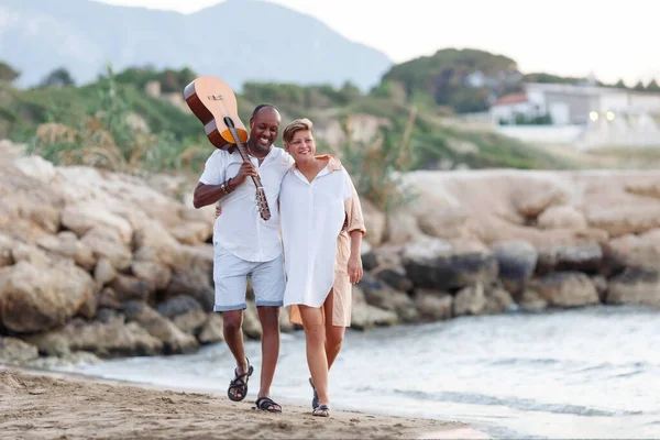 Feliz raça mista, casal de meia-idade abraçando enquanto caminhava na praia segurando uma guitarra. Homem segurando guitarra com mulher — Fotografia de Stock