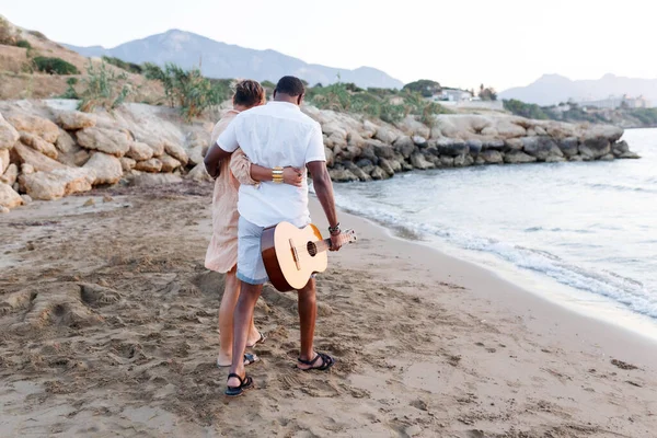 Feliz raça mista, casal de meia-idade abraçando enquanto caminhava na praia segurando uma guitarra. Homem segurando guitarra com mulher — Fotografia de Stock
