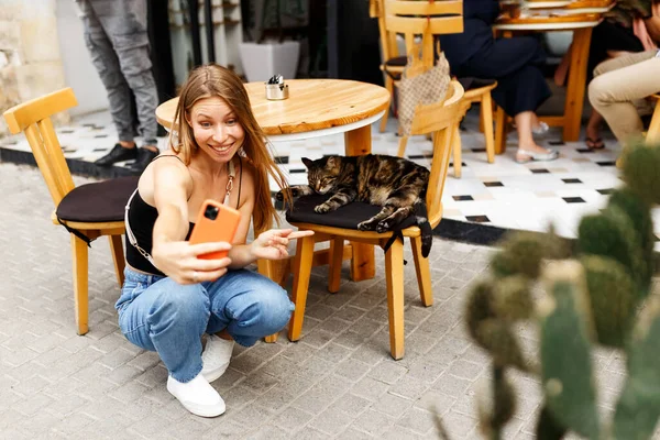 Porträt eines attraktiven, fröhlichen jungen Mädchens mit Smartphone, Blogger macht Selfie oder Live-Übertragung mit Katze in der Innenstadt im Freien Caféplatz — Stockfoto