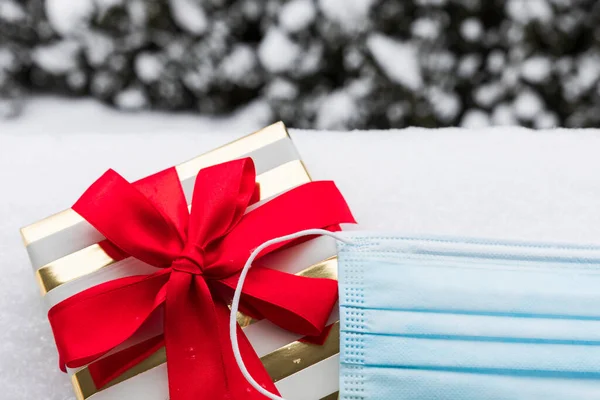 Covid-19 virüsünün tatillerin kutlanması üzerindeki etkisinin kavramsal fotoğrafı. Bir Noel hediyesi ve kar altında bir tıbbi maske..
