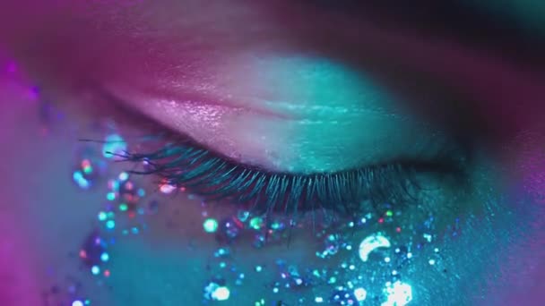 Ojos marrones femeninos en luz de neón. Joven chica sexy en un club nocturno. Mirada macro del ojo humano. Maquillaje de purpurina de moda en la cara. Brillantes destellos. Color rosa-azul-verde y nueva luz, euforia. — Vídeos de Stock