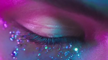 Neon ışıkta kahverengi gözlü bir kadın. Gece kulübündeki genç seksi kız. İnsan gözünün makro görüntüsü. Yüzünde modaya uygun sim makyajı var. Parlak parıltılar. Pembe-mavi-yeşil renk ve yeni ışık, coşku.