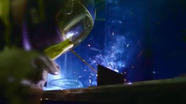 Fabrika işçisi metali kaynaklıyor. Adam kaynak yapıyor. Kaynak makinesi kullanarak argon veya elektrot ile kaynak yapmak. Metal yapılar üreten bir endüstriyel girişim. Kıvılcımlar ve parıltılar uçuşur. Yavaş çekim.