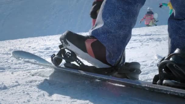 A lány feltesz egy téli snowboardot a hegyoldalon. A győzelem fogalma a sportban. Beszúrja a lábat, rögzíti a kötéseket a deszkán. Téli utazás a síközpontban — Stock videók