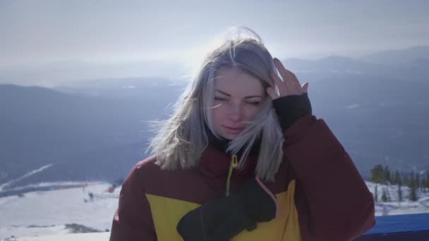 Porträtt: Vacker sexig tjej ryttare på en bakgrund av snöiga berg, tittar på kameran. Håret rör sig i vinden. Vintersemester i skid- och snowboardsorten. Långsamma rörelser. — Stockvideo