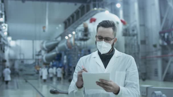 Wetenschapper of ingenieur met een medisch beschermend masker in een militaire hangar. Hij maakt notities in een tablet, maakt een ruimteraket klaar voor lancering tijdens een pandemie. Ruimtetechnologieën tijdens een pandemie — Stockvideo