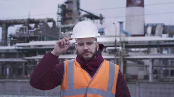 Retrato de un ingeniero profesional de la industria pesada en un sombrero blanco cerca de una fábrica mirando a la cámara. Trabaja en una refinería de petróleo o una fábrica de gas. Desarrollo de los yacimientos petrolíferos del norte. — Vídeos de Stock