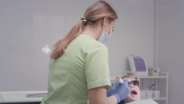 Una doctora encantadora trabaja con un paciente infantil. Comprueba el estado de los dientes de los niños. Doctor examinando los dientes de una adolescente en silla. La chica pone instrumentos médicos en la boca de los chicos — Vídeos de Stock