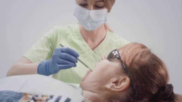 Красивая женщина-врач работает с пациентами постарше. Врач осматривает зубы пожилой женщины в кресле. Девушка кладет медицинские инструменты в рот бабушки. — стоковое видео