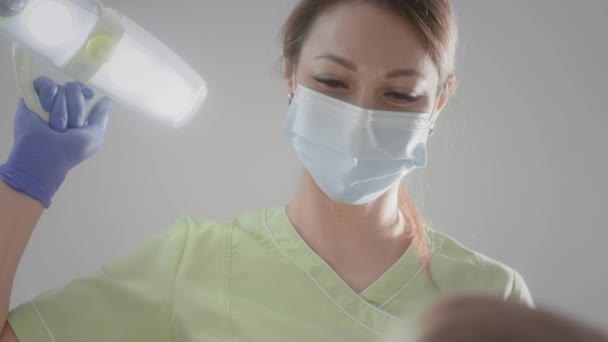 La encantadora doctora está tocando una bombilla médica. Trabaja con un paciente infantil. Comprueba el estado de los dientes de los niños. Ella pone instrumentos médicos en la boca de los chicos. Odontología infantil. — Vídeo de stock