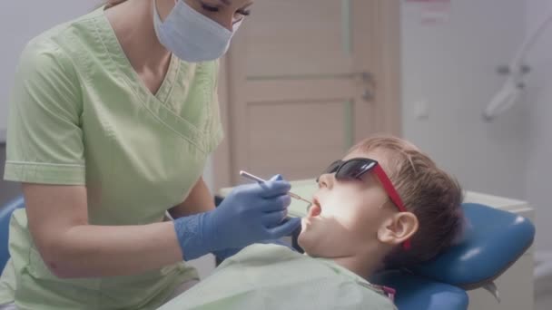 Uma médica encantadora está a tocar numa lâmpada médica. Trabalha com uma criança paciente. Verifica a condição dos dentes das crianças. Ela põe instrumentos médicos na boca dos rapazes. Odontologia infantil. — Vídeo de Stock