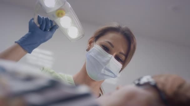 Uma médica adorável trabalha com uma paciente idosa. Move uma lâmpada de luz. Doutor examinando dentes de mulher idosa na cadeira. A menina coloca instrumentos médicos em sua boca avós — Vídeo de Stock