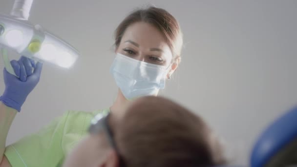 La encantadora doctora está tocando una bombilla médica. Trabaja con un paciente infantil. Comprueba el estado de los dientes de los niños. Ella pone instrumentos médicos en la boca de los chicos. Odontología infantil. — Vídeo de stock