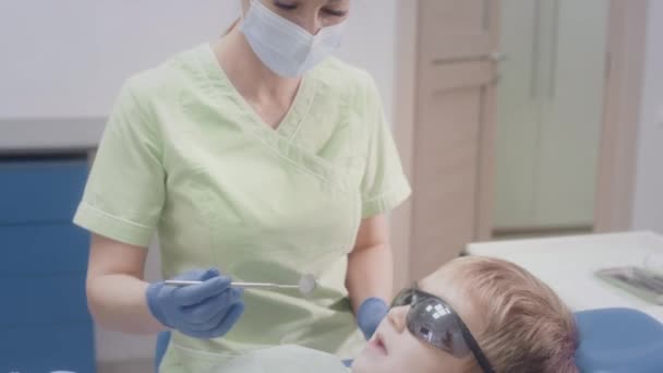 Urocza lekarka pracuje z dzieckiem. Sprawdza stan dziecięcych zębów. Lekarz badający zęby nastolatki na krześle. Dziewczyna wkłada instrumenty medyczne do ust chłopców. — Wideo stockowe