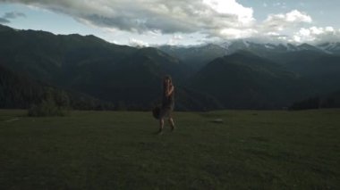 Genç güzel kız gün batımında dağlarda yeşil çimlere basıyor. Gezgin kıyafetleri, uzun elbiseler ve şapkalar. Hayatın tadını çıkarıyor, gülümsüyor. Karla kaplı dağlar.