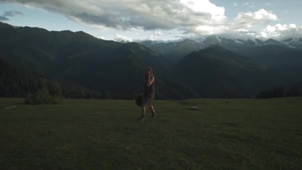 Joven hermosa niña camina sobre hierba verde en las montañas durante la puesta del sol. Ropa de viajero, vestido largo y sombrero. Disfrutando de la vida, sonriendo. Montañas cubiertas de nieve. — Vídeos de Stock